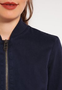 Námořnická bombardovací bunda s vroubkovaným límcem a kovovým zipem, vyrobená z hladké tkaniny s jemným leskem.