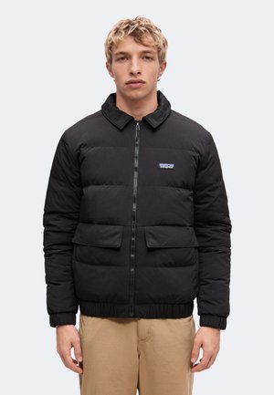 Junger Mann mit kurzen blonden Haaren, der eine schwarze Steppjacke mit Fronttaschen und beigen Hosen trägt, vor einem schlichten Hintergrund stehend.