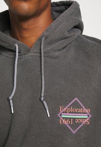 Tmavě šedá mikina s kapucí se šňůrkami, s diamantovým logem v růžové a zelené barvě s nápisem "Exploration Since 1993." Jemná textura látky.