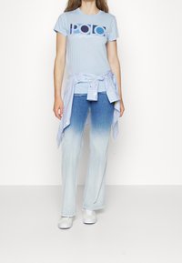 Světle modré polo tričko s výrazným logem a bílým textem, spárované se světle modrými džínami a pruhovanou košilí uvázanou v pase. Bílé tenisky.