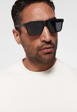Uomo con capelli corti e scuri e barba, che indossa occhiali da sole neri quadrati e una camicia bianca semplice su uno sfondo chiaro.