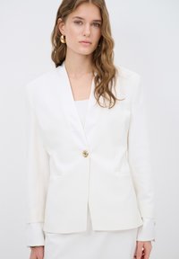 White blazer with a tailored design, single gold button, and two front pockets. Smooth texture and shawl lapels.