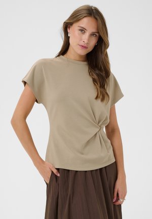 Femme aux longs cheveux bruns portant un haut beige à manches courtes avec un détail torsadé à l'avant et une jupe marron foncé, debout avec une main dans la poche.
