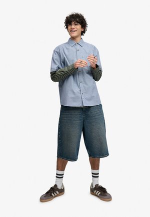 Ung mand med krøllet hår iført lagvise lange og korte ærmer, lange denimshorts, stribede sokker og sneakers, smilende med hænder midt i en gestus.