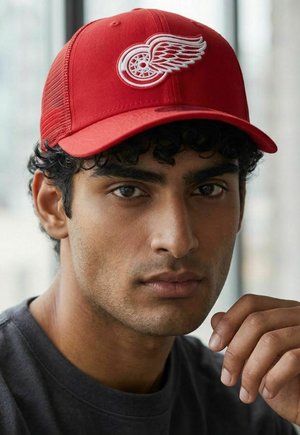 Young man with curly hair wearing a red cap with a white winged wheel logo, resting his hand near his face, looking forward.