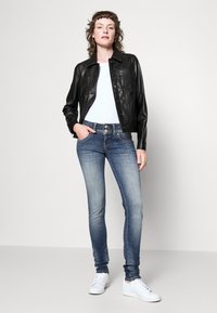 Mujer de pie vistiendo una chaqueta de cuero negra con botones, una camiseta blanca, jeans ajustados de mezclilla azul deslavada con doble botón y zapatillas blancas, con las manos en los bolsillos.