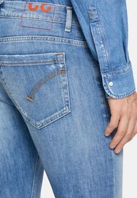 Wyczerpane niebieskie dżinsy z pomarańczowymi przeszyciami. Posiadają prostokątną tylną kieszeń oraz widoczną etykietę z logo powyżej pasa.