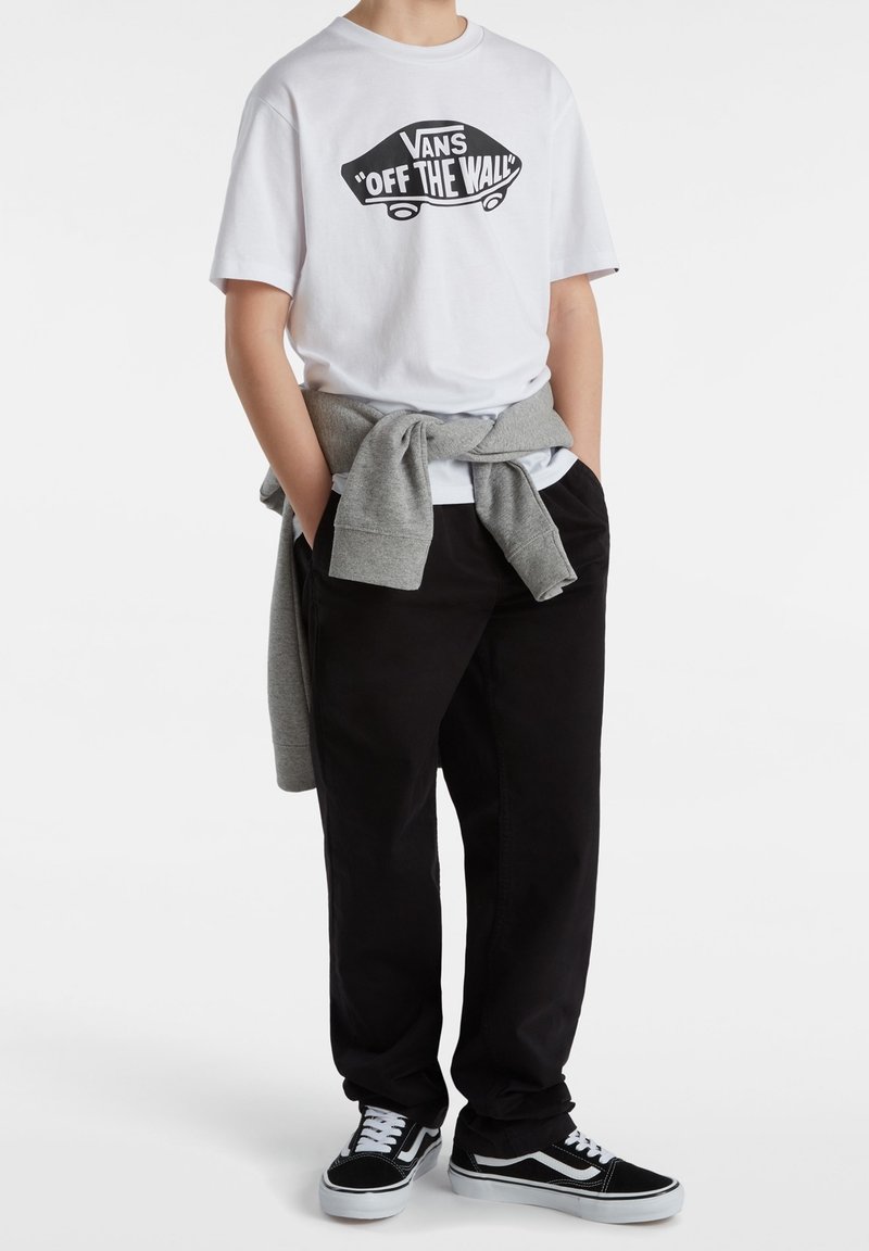 Personne portant un T-shirt blanc Vans « Off The Wall », un pantalon noir, des baskets Vans noires et un sweat gris noué autour de la taille, debout les mains dans les poches.