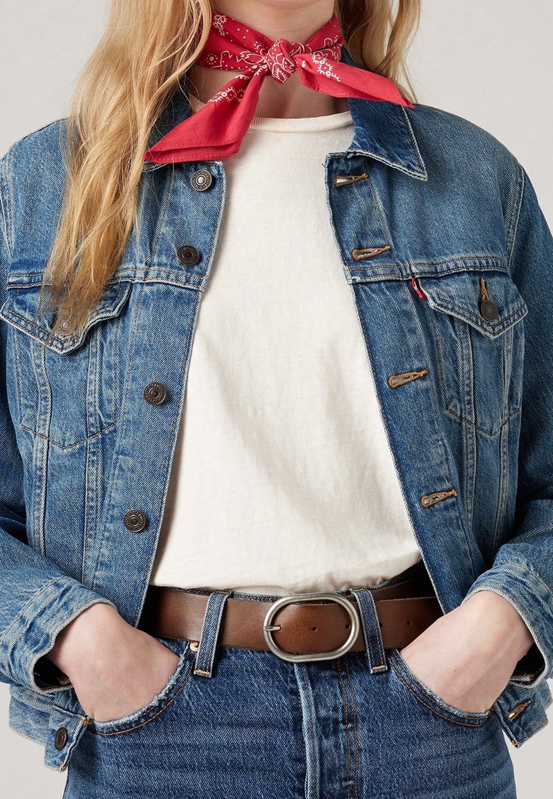 Chaqueta de mezclilla de color azul claro con cierres de botón y bolsillos en el pecho, llevada sobre una camisa blanca. Cinturón de cuero marrón con una hebilla de plata. Pañuelo rojo atado alrededor del cuello.