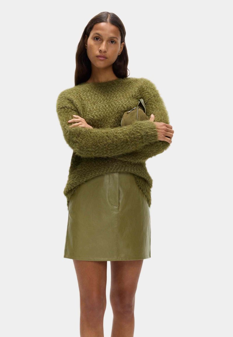 Pullover verde peloso con una vestibilità ampia, abbinato a una gonna corta in pelle oliva aderente. La modella tiene una piccola borsa verde.
