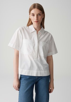 Una camicia bianca a maniche corte con colletto, dotata di due tasche anteriori, taglio dritto e texture di cotone morbido. Indossata con jeans a gamba larga blu.