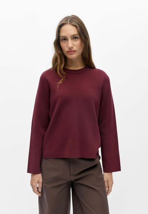 Femme aux longs cheveux ondulés porte un pull bordeaux à manches longues et un pantalon marron sur un fond blanc uni.