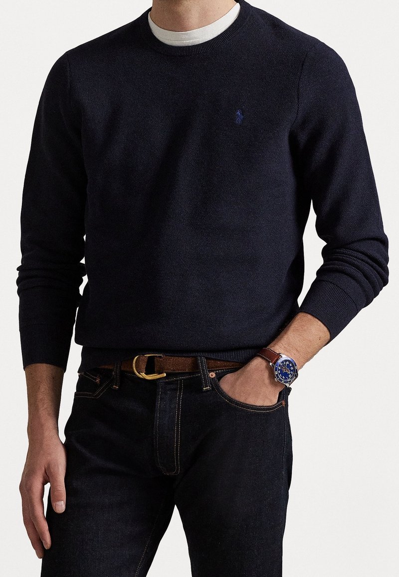 Homme portant un pull col rond bleu marine foncé par-dessus une chemise blanche, un jean foncé avec une ceinture marron, et une montre-bracelet à cadran bleu avec un bracelet en cuir marron.