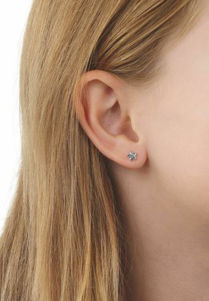 Zbliżenie ucha osoby z długimi blond włosami, noszącej mały, srebrny kolczyk w kształcie gwiazdy z osadzonymi kamieniami.