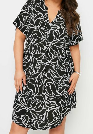 Kobieta w rozmiarze plus nosząca czarno-białą sukienkę o krótkim rękawie z abstrakcyjnym kwiatowym wzorem, dekoltem w szpic i srebrną bransoletką.
