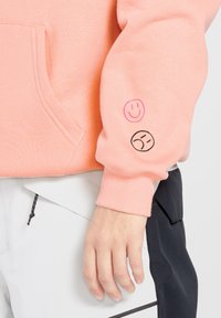 Helle rosa Kapuzenjacke aus weichem Stoff mit einer Tasche auf der Vorderseite. Der Ärmel zeigt ein pinkes Smiley-Gesicht und ein schwarzes trauriges Gesicht.
