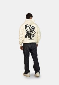 Felpa over-size color crema con grafica nera "RISE & SHINE" sul retro, abbinata a jeans scuri e sneakers di colore chiaro.