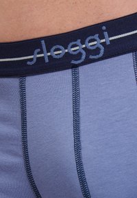 Slip da uomo in tessuto azzurro chiaro con elastico blu scuro. Presenta cuciture visibili e logo. Texture morbida, design aderente.
