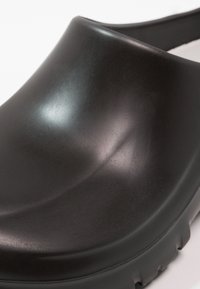 Schwarzer Gummischuh mit einem glatten, geschwungenen Design, runder Zehenpartie und einer strukturierten Sohle für besseren Grip, ideal für einfaches Hineinschlüpfen.