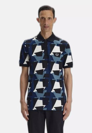 Man staat en draagt een polo met korte mouwen met een geometrisch patroon in blauw, zwart en wit, samen met een zwarte broek, tegen een effen achtergrond.