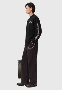 Camisa negra de manga larga con estampado gráfico en el pecho y diseños rojos a lo largo de las mangas, combinada con pantalones anchos y oscuros y botas negras.