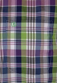 Rutigt skjorta med lila, grön, blå och vita rutor. Inkluderar en liten ficka med logotyp och vit sömdekoration.