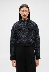 Fiatal nő fekete-kék texturált, rövidített dzsekit visel, nagyméretű lehajtható zsebekkel, egyszerű fehér háttér előtt áll.