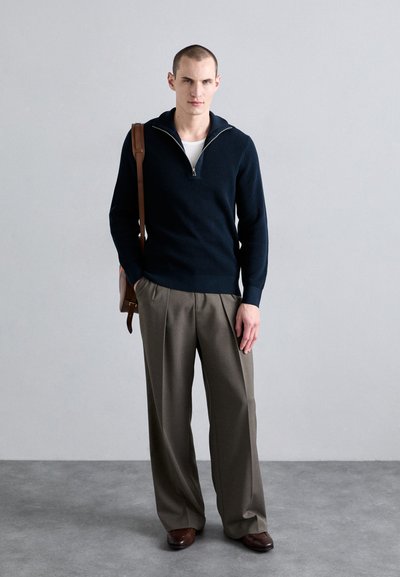 Marineblauer gerippter Strickpullover mit Viertel-Reißverschluss, darunter ein cremefarbenes Hemd, kombiniert mit mittelgrauen, gefalteten Hosen und braunen Lederschuhen, während er eine braune Tasche hält.