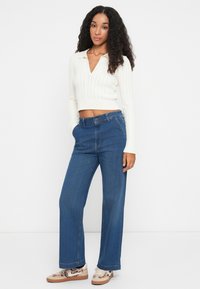 Maglione corto bianco a maglia con motivo a treccia, abbinato a jeans a vita alta blu con gamba larga e sneaker con motivo.