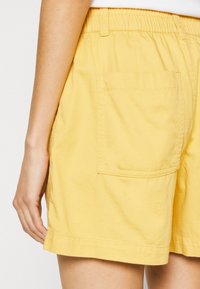 Närbild av en person i gula elastiska midjebyxor med en stor bakficka och en vit topp.