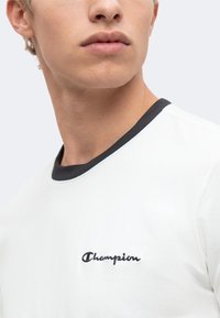 Νέος άνθρωπος που φοράει λευκό t-shirt με μαύρη επένδυση και τη λέξη champion κεντημένη στο στήθος, κοιτάζοντας ελαφρά στο πλάι.