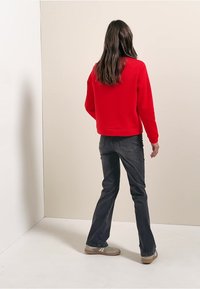 Červená mikina s jednoduchým designem, kombinovaná s tmavými džíny a světlými botami. Materiál vypadá měkký a střih je volný.