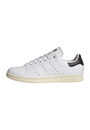 Valge Adidas Stan Smith toss, millel on läbi torgatud triibud ja tumedat pruunikat tekstuuriga kannaosa, kreemjas tall ja valged paelad, vaadatuna küljelt.