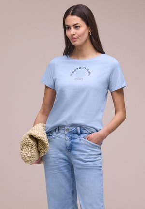 Kvinde iført lyseblå T-shirt med teksten "Summer is my Season" og jeans, holder en beige flettet taske med den ene hånd i lommen, neutral baggrund.