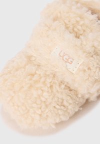 Beige koselige tøfler laget av fluffy shearling-materiale, med en liten UGG-logo. Myk tekstur med rund form.