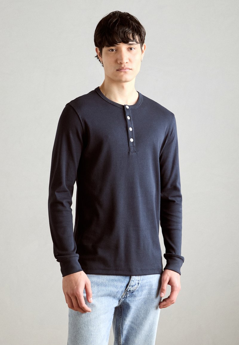 Marineblå langermet henley-skjorte i myk stoff, med rund krage og fem-knapps placket, brukt sammen med lyseblå jeans.
