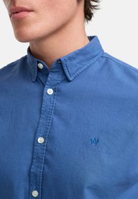 Blå bomullsskjorte med nedbrettbar krage, med hvite knapper og en liten brodert logo i det nedre hjørnet. Glatt tekstur.