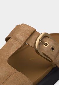 Κοντινή λήψη στο λουράκι σανδαλιού από ταμπά σουέντ με χρυσή αγκράφα και λεπτομέρειες ραφής πάνω σε καφέ πάτο και μαύρη σόλα.