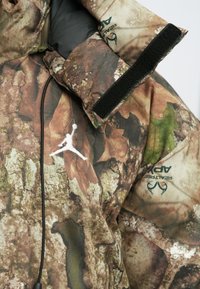 Giacca mimetica che presenta una combinazione di texture marroni, verdi e sabbia che ricordano la corteccia degli alberi e le foglie, con un logo bianco sul davanti.