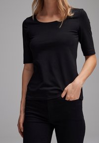 Woman wearing a black fitted half-sleeve top and black pants, with one hand in her pants pocket, against a plain gray background.