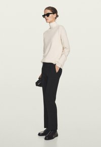 Donna che indossa un maglione bianco a collo alto, pantaloni neri a sigaretta, mocassini neri e occhiali da sole, mentre tiene una piccola borsa nera.