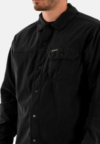 Zwarte corduroy shirt met een knoopsluiting aan de voorkant, een gespreide kraag en twee borstzakken met drukknoopsluitingen. De textuur van de stof zorgt voor een ribbelige uitstraling.