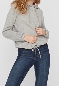 Sudadera con capucha gris con puños elásticos y cintura ajustable con cordón, combinada con jeans ajustados de color azul oscuro. Tejido suave y diseño informal.