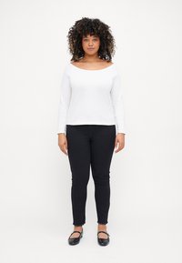 Top blanco de manga larga y hombros al aire, combinado con pantalones skinny negros. Los pantalones tienen dobladillos deshilachados y ella lleva zapatos planos negros.
