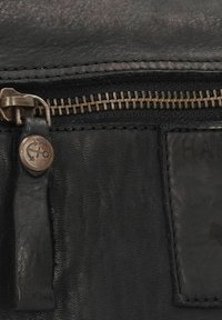 Detail einer schwarzen Lederhandtasche mit einem Metallreißverschluss, einem Lederanhänger mit Ankeremblem und einer strukturierten Oberfläche.