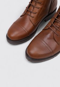 Botas marrones de cuero hasta el tobillo con punteras redondas, cordones, detalles de costura y suela de goma texturizada. Presentan un acabado suave y un brillo sutil.