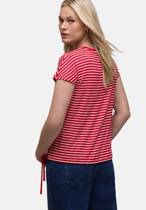Femme blonde tournant le dos, portant un t-shirt à manches courtes rayé rouge et blanc et un jean bleu foncé, sur fond uni.