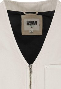 Gros plan sur un vêtement beige avec une fermeture éclair argentée, une doublure intérieure noire et une étiquette indiquant Urban Classics Organic, taille M, fabriqué au Pakistan.