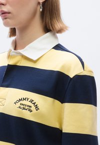 Κίτρινο και ναυτικό ριγέ πόλο πουκ�άμισο με λευκό γιακά, που διαθέτει κεντημένο λογότυπο "TOMMY JEANS" και υφασμάτινη υφή.