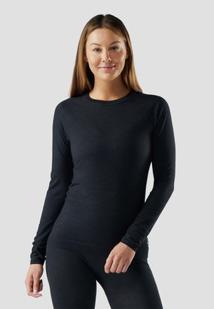 Žena v těsně přiléhajícím černém dlouhém tričku s rukávy a odpovídajících černých legínách, usmívající se a stojící na jednoduchém světle šedém pozadí.